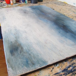 Op de afbeelding: Een close-up van een wit canvas met een blauwe en grijze gradi&euml;nt. Het canvas ligt op een houten oppervlak met verfspatten.