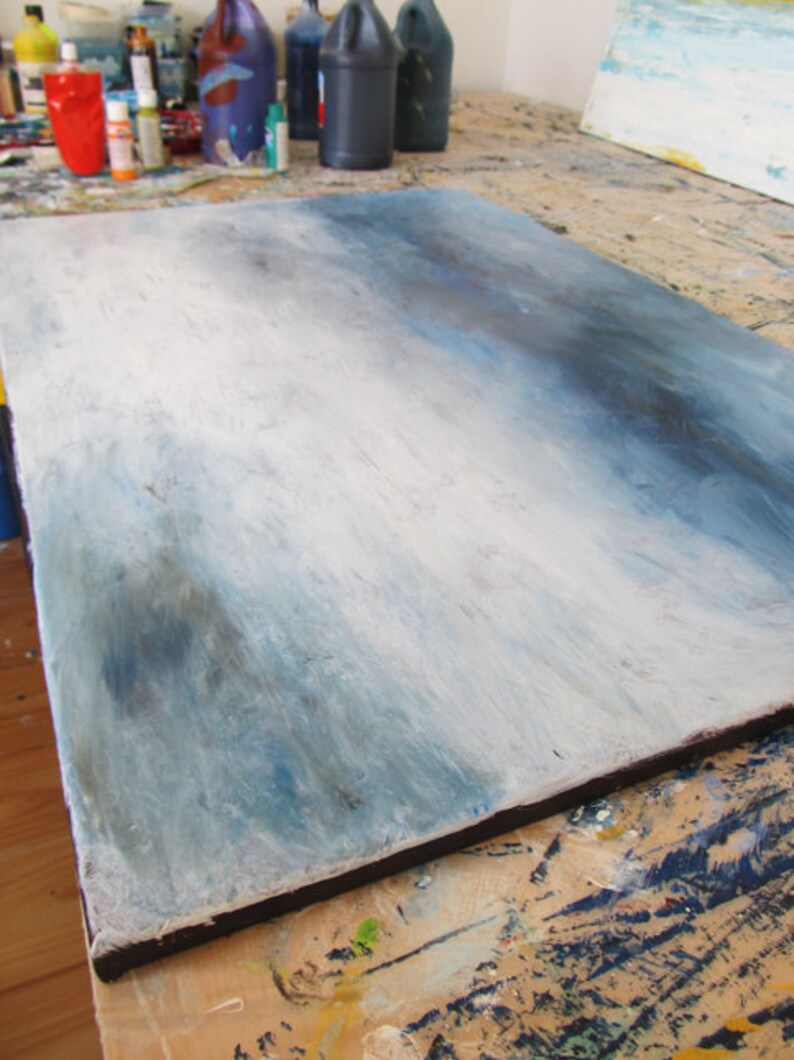 Op de afbeelding: Een close-up van een wit canvas met een blauwe en grijze gradi&euml;nt. Het canvas ligt op een houten oppervlak met verfspatten.