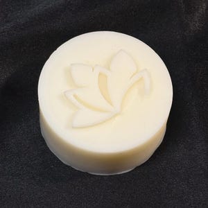May include: A round, off-white bar of soap featuring a raised, embossed lotus flower design on its top surface. The soap has a smooth, solid texture and a clean, circular shape, presenting a simple and uniform appearance throughout the entire piece.