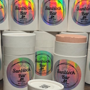 May include: White cylindrical containers with holographic labels that read "Sunblock Bar" and "Tallow Glow Approx SPF 20". One container is open, revealing a pink sunscreen stick. The containers are arranged in a group.