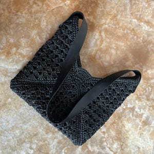 May include: Black crochet tote bag with a geometric pattern. The bag has a rectangular shape with a wide, black strap. The bag is made of a woven material and is suitable for carrying items.