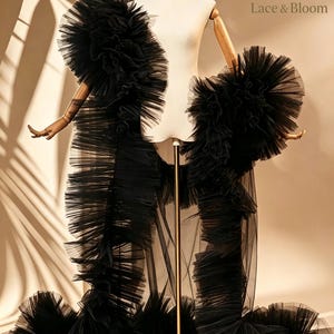Pode incluir: Um roupão de tule preto transparente com babados volumosos ao longo das bordas e mangas. A peça é exibida em um manequim e inclui o texto Lace & Bloom no canto superior direito. O design consiste em camadas de tecido plissado.