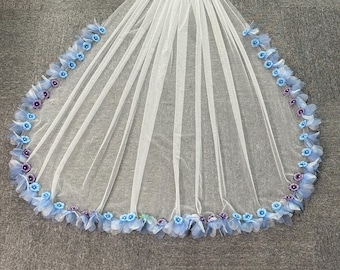 May include: A white tulle bridal veil with a cascade design. The veil is adorned with light blue and purple fabric flowers along the edge and at the top. The veil is spread out on a gray surface.