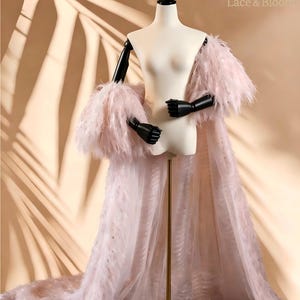 May include: A floor-length pale pink robe featuring long sleeves adorned with dense feathers. The garment is crafted from a sheer, textured fabric with a subtle wavy pattern throughout. The robe is displayed on a mannequin with black arms.