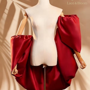 May include: A red satin gown with dramatic, oversized sleeves and a high-low hemline is displayed on a mannequin. The dress features a fitted bodice and a flowing skirt. The text "Lace & Bloom" is visible on the upper right.