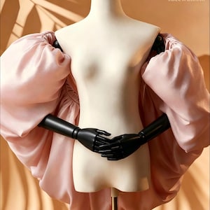 Puede incluir: Una prenda de color rosa pálido con mangas abullonadas voluminosas y de gran tamaño hechas de una tela suave similar al satén. La pieza se muestra en un maniquí con brazos negros articulados. El texto 'Lace & Bloom' aparece arriba a la derecha.