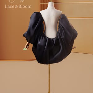 May include: A black dress with voluminous sleeves and a gathered skirt is displayed on a gold dress form. The dress is made of a shiny fabric and is set against a warm, neutral background. The text "Lace & Bloom" is visible in the upper left corner.