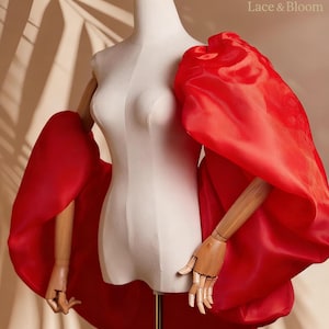 May include: A white dress form with wooden arms and hands is draped with a vibrant red, voluminous fabric. The fabric creates large, dramatic sleeves. The dress form is on a gold stand, with the text "Lace & Bloom" visible.