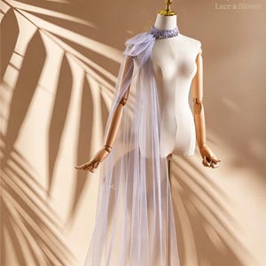May include: A light purple tulle cape draped over a mannequin. The cape features a beaded choker and a shoulder accent. The mannequin is on a gold stand, with the text "Lace & Bloom" in the upper right corner.