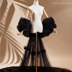 Może przedstawiać: Czarna tiulowa suknia ślubna z długim trenem i rękawami wykończonymi piórami jest prezentowana na manekinie. Suknia ma dopasowany gorset i zwiewną spódnicę. Manekin stoi na złotym stojaku, a tło jest w neutralnym kolorze.