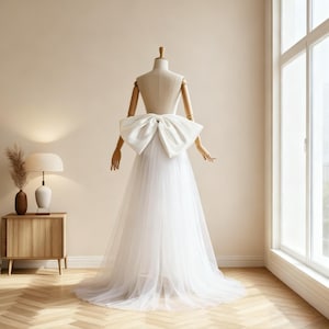 May include: A white wedding dress on a mannequin. The dress features a large, satin bow at the waist and a flowing tulle skirt. The mannequin is positioned in front of a window, with a neutral-toned wall and wooden furniture in the background.