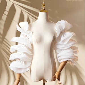 May include: A white dress form with layered, ruffled sleeves. The sleeves are made of a sheer fabric and are attached to wooden articulated arms. The dress form is on a metal stand. The logo "Lace & Bloom" is visible in the upper right corner.