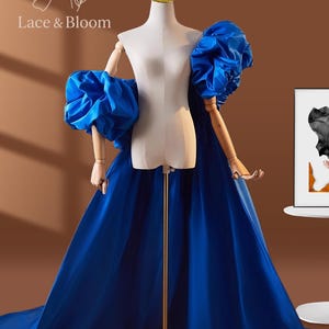May include: A royal blue gown with voluminous, puffed sleeves is displayed on a mannequin. The dress features a long, flowing train and is set against a neutral background. The brand name "Lace & Bloom" is visible in the upper left corner.
