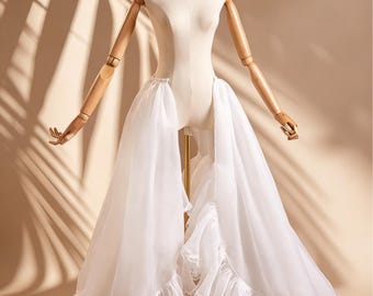May include: A white sheer overskirt featuring a high-low design with voluminous ruffled edges and delicate lace trim. The garment is displayed on a mannequin and includes the text Lace & Bloom in the upper right corner of the image.
