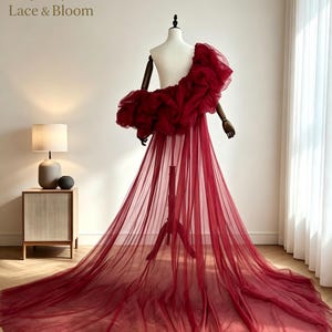 Puede incluir: Un vestido rojo oscuro de un solo hombro con un corpiño de tul voluminoso y con volantes, y una falda larga y fluida de tul transparente que llega hasta el suelo. La prenda se exhibe en un maniquí, destacando la tela en capas.