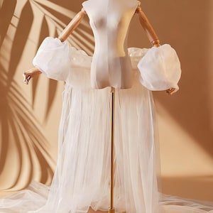 Puede incluir: Un maniquí de costura con brazos de madera y un soporte dorado, cubierto con una falda de tul blanco y mangas abullonadas transparentes. El maniquí es de color neutro, sobre un fondo beige cálido, creando una estética suave y elegante.