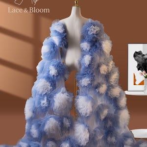 May include: A blue and white tulle bridal robe on a mannequin. The robe is adorned with layers of ruffled tulle, creating a cloud-like effect. The text "Lace & Bloom" is visible in the upper left corner. The robe is a light blue color with white accents.