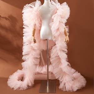 May include: A light pink tulle robe with ruffled trim, displayed on a mannequin. The robe features a long train and a cascading design. The mannequin has wooden arms and is set against a brown backdrop.