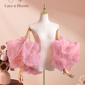 Puede incluir: Un par de mangas de tul rosa en un maniquí. Las mangas están hechas de capas de tela suave y transparente, creando un efecto voluminoso. El maniquí está sobre un soporte dorado, con el logo "Lace & Bloom" visible.