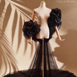 May include: A black evening gown on a mannequin. The gown features large, ruffled sleeves and a flowing tulle skirt that extends into a train. The mannequin is set against a neutral background with the text "Lace & Bloom".