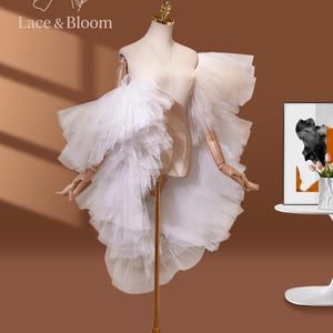 May include: A white tulle dress displayed on a mannequin. The dress features layered ruffles and a high-low hemline. The mannequin has wooden arms and a gold-colored stand. The logo "Lace & Bloom" is visible in the upper left corner.