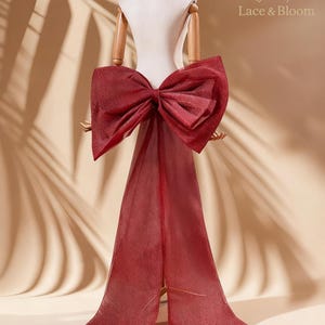 May include: A burgundy bow and train are displayed on a dress form. The bow is large and textured, with a shimmering finish. The train flows down, creating a dramatic effect. The text "Lace & Bloom" is visible in the upper right corner.