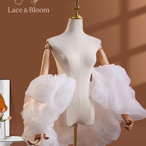 Puede incluir: Un maniquí blanco con brazos de madera y cuello dorado. El maniquí está adornado con una tela blanca transparente y voluminosa, creando un efecto de capas. El texto "Lace & Bloom" es visible en la esquina superior izquierda.
