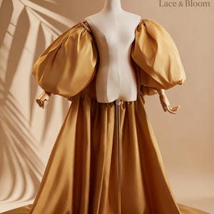 Könnte beinhalten: Ein goldgelbes Kleid mit großen Puffärmeln und einem fließenden Rock wird auf einer Schaufensterpuppe präsentiert. Das Kleid hat eine dramatische Silhouette mit einem eng anliegenden Oberteil und einer langen Schleppe. Auf dem neutralen Hintergrund steht "Lace & Bloom".