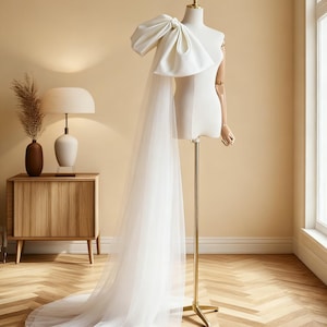 Puede incluir: Vestido de novia blanco con un gran lazo en el hombro, expuesto en un maniquí. El vestido presenta una cola fluida y transparente. El maniquí está sobre un soporte dorado, en un interior de tonos neutros.