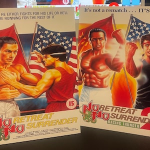 May include: Two video game box covers for the game No Retreat No Surrender 2: Raging Thunder. The covers feature two different scenes of martial arts fighting. The first cover features two men in red, white, and blue fighting in a boxing ring. The second cover features two men in red, white, and blue fighting in a street setting. Both covers feature the game's title and logo.