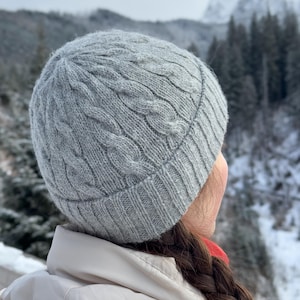 May include: A grey knitted hat with a cable knit pattern. The hat is being worn by a person with long brown hair, standing in front of a snowy mountain landscape.