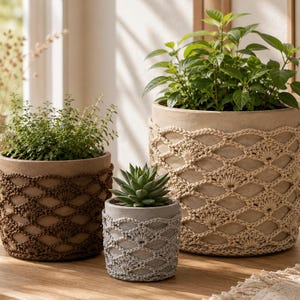 Può includere: Tre vasi cilindrici di diverse dimensioni, ciascuno rivestito da una copertura decorativa all'uncinetto. Il vaso più grande presenta un motivo all'uncinetto color crema, quello medio una copertura marrone scuro e il più piccolo una grigia.