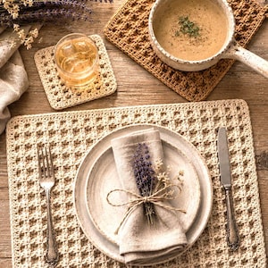 May include: A cream-coloured crocheted rectangular placemat and square coaster, a tan crocheted square trivet, a ceramic bowl with soup, a ceramic plate set with a linen napkin tied with twine and lavender, a silver fork, and a silver knife.