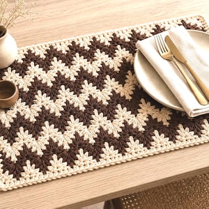 May include: A rectangular crochet placemat featuring a repeating chevron pattern in alternating dark brown and cream yarn. The placemat has a thick, textured border and is crafted with a dense stitch, creating a consistent geometric design throughout.