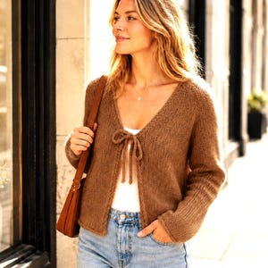 May include: A brown long-sleeved knit cardigan with a V-neckline and a front tie closure. The garment is worn over a white top and paired with light blue denim jeans, accompanied by a brown leather shoulder bag with a thin strap.