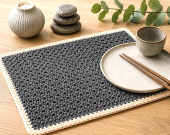 May include: A rectangular crochet placemat featuring a dark charcoal gray center with a textured star-like stitch pattern, bordered by a thick, light cream-colored crocheted edge. The placemat is flat and rectangular with a consistent yarn texture.
