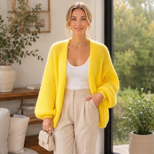 May include: A bright yellow open-front knit cardigan with long, loose sleeves, worn over a plain white tank top and beige high-waisted trousers. A small, structured cream-colored handbag with a gold clasp is held in one hand.