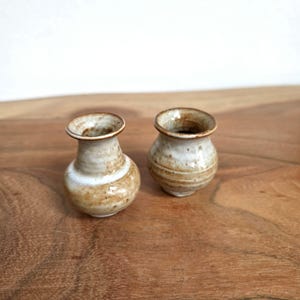 Könnte beinhalten: Zwei kleine, handgefertigte Keramikvasen mit bauchigem Körper und ausgestelltem Rand. Die Keramik weist eine gesprenkelte, cremeweiße Glasur mit warmen braunen und beigen Erdtönen auf, die ein glattes, glänzendes Finish und eine handgetöpferte Optik zeigt.