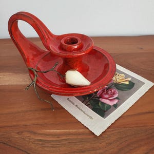 Könnte beinhalten: Ein leuchtend roter Kerzenhalter aus Keramik mit Griff, der einen weißen Gegenstand und einen kleinen Zweig hält. Eine Vintage-Postkarte mit Rosenillustrationen liegt daneben. Der Kerzenhalter steht auf einer Holzoberfläche.