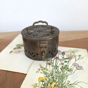 Könnte beinhalten: Kleine, runde, antike Messingbox mit Griff und Verschluss. Deckel und Seiten sind mit kreuzförmigen Aussparungen verziert. Die Box liegt auf einem Vintage-Botanikausdruck mit bunten Blumen.
