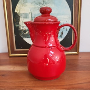 Könnte beinhalten: Eine glänzend rote Keramik-Kaffeekanne mit Deckel und Griff. Die Kanne hat einen abgerundeten Körper und einen Ausguss zum Ausgießen. Der Hintergrund enthält ein gerahmtes Bild mit einer Winterszene.