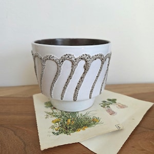 May include: White ceramic planter with a textured, wavy brown design around the exterior. The interior is dark brown. The planter sits on a small base. A botanical print is visible beneath the planter.