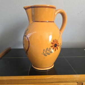 Op de afbeelding: Een gele keramische kan met een handvat en tuit. De kan is versierd met een bloemenontwerp in bruin, oranje en goud. De kan staat op een zwart oppervlak.