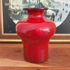 Könnte beinhalten: Eine leuchtend rote Glasvase mit ausgestelltem Boden und schmalem Hals. Die Vase hat eine glänzende Oberfläche und einen dunkelroten Rand. Der Hintergrund zeigt ein gerahmtes Gemälde mit einer Winterszene.