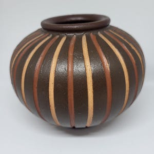 Könnte beinhalten: Eine runde, braune Keramikvase mit vertikalen Streifen in Braun- und Beigetönen. Die Vase hat einen dunkelbraunen Rand und eine strukturierte Oberfläche. Diese dekorative Vase eignet sich für die Inneneinrichtung.