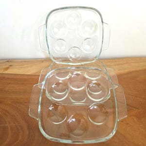 May include: Two clear glass escargot dishes stacked on a wooden surface. Each dish is square with rounded indentations for the snails and handles on the sides. The dishes are empty and ready for use.