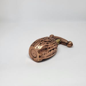Könnte beinhalten: Ein kupferfarbener Flaschenverschluss mit einem strukturierten, oval geformten Körper und einem Griff. Der Verschluss hat eine metallische Oberfläche und ist für eine Flasche konzipiert. Der Griff hat eine zylindrische Form.