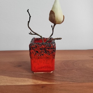 Könnte beinhalten: Eine kleine, quadratische, rote Keramikvase mit einem strukturierten schwarzen Rand. Ein Zweig mit einer cremefarbenen und braunen Blütenknospe befindet sich in der Vase. Die Vase steht auf einer Holzoberfläche.