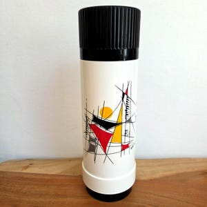 May include: A cylindrical white thermos with a black ribbed screw-top lid and a black base. The body features a graphic print of intersecting black lines, solid red and yellow triangles, and a yellow circle, creating an abstract geometric pattern.