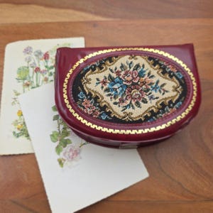Könnte beinhalten: Eine bordeauxfarbene Leder-Geldbörse mit einem ovalen Wandteppichmuster mit einem Blumenmuster in Rosa, Blau und Beige. Zwei Vintage-Blumenpostkarten liegen neben der Geldbörse auf einer Holzoberfläche.